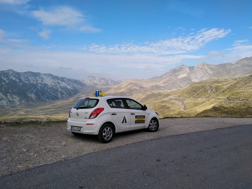 Az Apró Autósiskola fehér Hyundai i20 oktatóautója egy hegyi út mellett